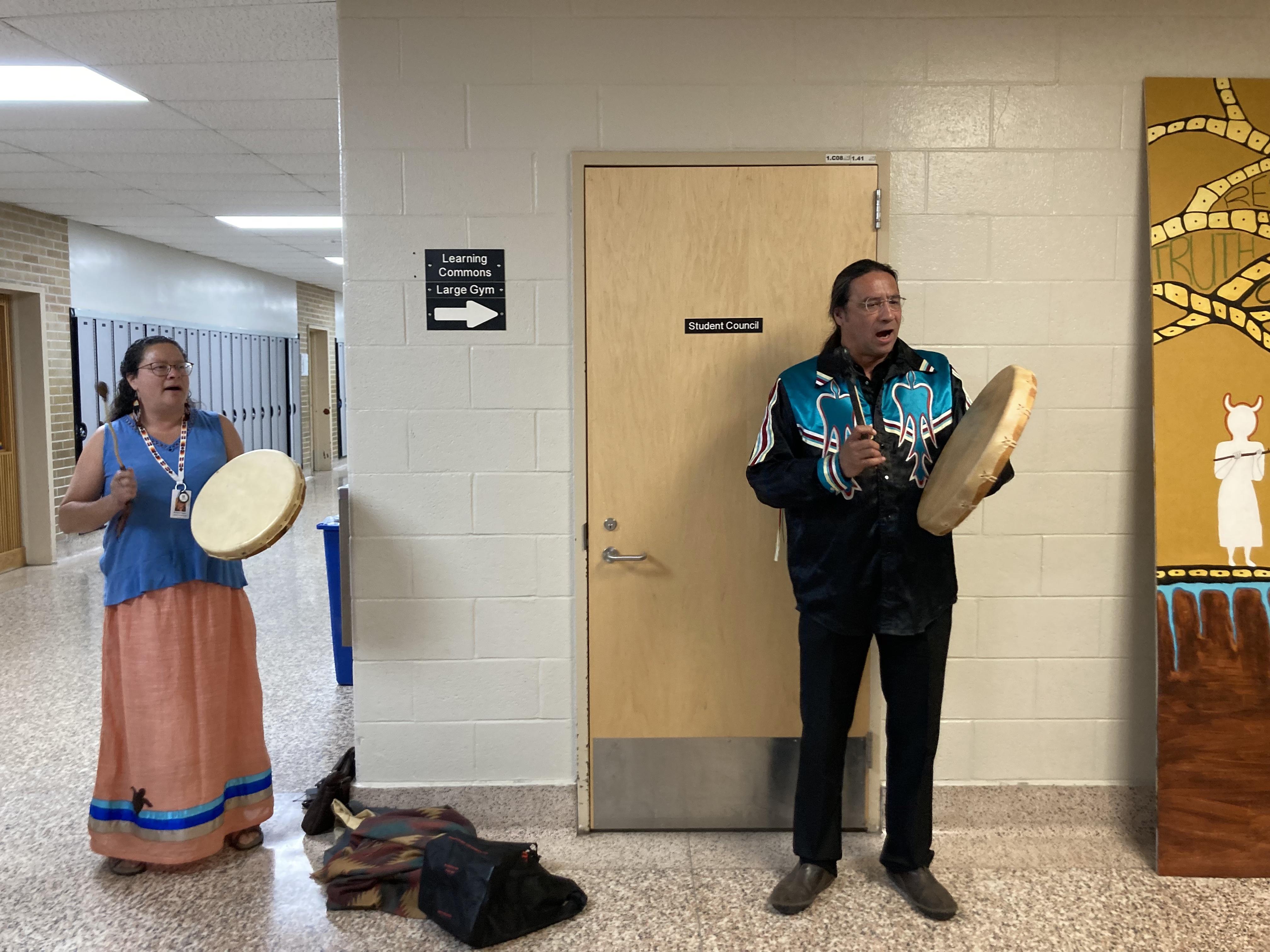 Monique Pregent and Christin Dennis play Indigenous drums in hallway beside Indigenous-inspired art piece